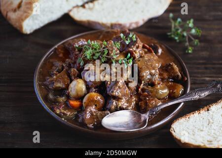 Ciotola di manzo Bourguignon guarnita con timo di limone fresco e servita con pane artigianale fatto in casa su un rustico tavolo di legno. Foto Stock