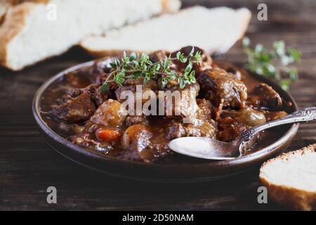 Ciotola di manzo Bourguignon guarnita con timo di limone fresco e servita con pane artigianale fatto in casa su un rustico tavolo di legno. Foto Stock