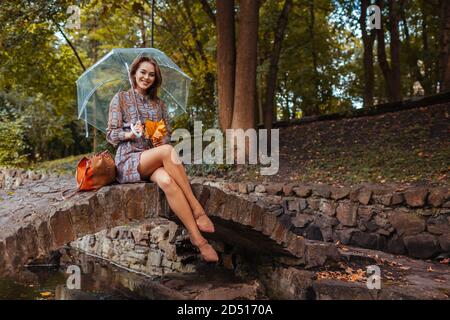 Felice giovane donna seduta sul margine da stagno in autunno parco sotto ombrello trasparente durante la pioggia. Passeggiata nella stagione autunnale Foto Stock