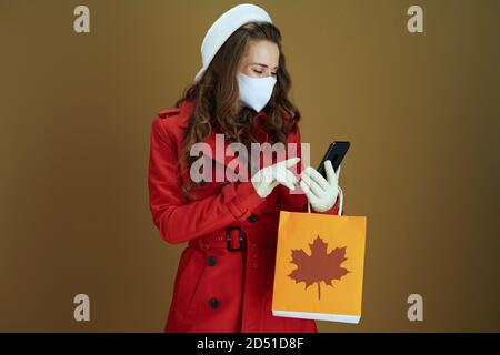 Vita durante la pandemia di covid-19. Casalinga elegante in cappotto rosso con borsa di carta per la spesa utilizzando applicazioni smartphone su sfondo beige. Foto Stock