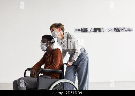 Ritratto minimo di afroamericano con sedia a rotelle e guardando i dipinti in galleria d'arte moderna con giovane donna che lo aiuta, entrambi indossando maschere, spazio di copia Foto Stock