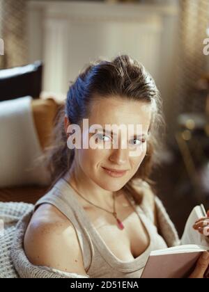 felice giovane donna in confortevole cardigan lavorato a maglia con libro nel moderno soggiorno in sole giornate autunnali. Foto Stock