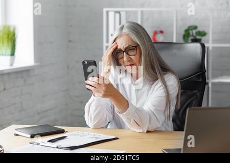 Confuso senior grigio-capelli bella donna che tiene smartphone. Donna d'affari imbarazzata ha problemi con rotto non funzionante telefono cellulare. Ricezione Foto Stock