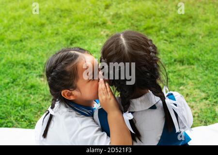 Due giovani ragazze whispering e condividere un segreto durante la sessione di giochi sul verde sullo sfondo di vetro Foto Stock