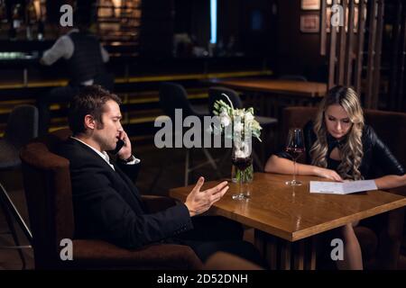 un giovane uomo d'affari risolve i problemi di lavoro in un ristorante al telefono, il suo compagno sceglie i piatti dal menu. Foto Stock