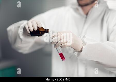 Un chimico maschio tiene in mano una provetta di vetro con un po' di liquido rosa e versa in essa un nuovo reagente. Foto Stock