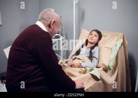 Un anziano sta raccontando a un bambino una storia in un ospedale, mentre il bambino sta rimanendo a letto e sta ascoltando con interesse. Foto Stock