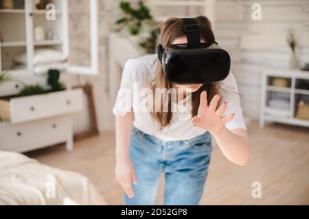 Giovane donna in jeans e T-shirt bianca che indossa casco di realtà virtuale gioca a gioco, opere, orologi video nella stanza accanto al letto. Concetto di tecnologia moderna Foto Stock