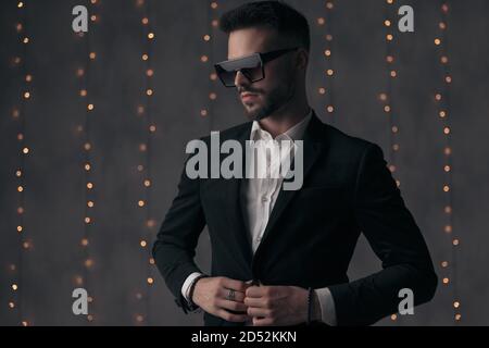 Modello di moda sicuro chiudendo la giacca e guardando via mentre in piedi su sfondo grigio studio con luci Foto Stock