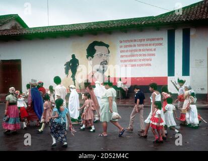 La gente in una sfilata di festival passa un murale rivoluzionario Sandjnista con l'immagine del leader caduto, Carlos Fonseca, e citazione da lui, a Granada, Nicaragua nel periodo Sandinista, 1986 Foto Stock