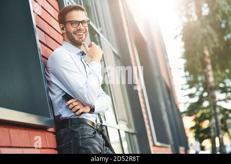 Giovane uomo d'affari felice che indossa auricolari wireless che tengono lo smartphone, guardando la fotocamera e sorridendo mentre si sta in piedi all'aperto Foto Stock