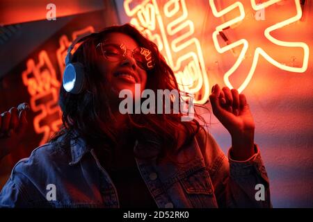 Donna afroamericana felice indossando le cuffie che ascoltano la musica nella luce del neon. Foto Stock