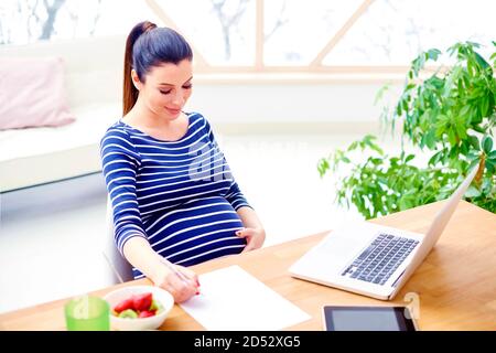 Fiducioso giovane donna incinta seduta nella parte anteriore del computer portatile e e facendo alcuni documenti mentre si lavora da casa. Foto Stock