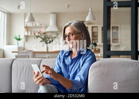 Donna anziana matura che usa uno smartphone seduto sul divano a casa. Foto Stock