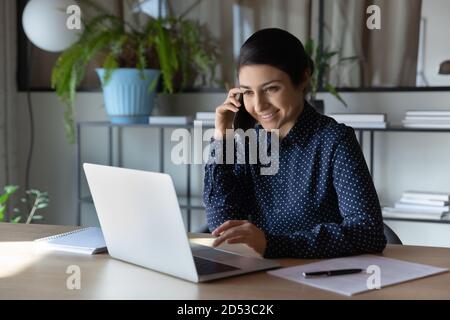 Sorridente donna indiana lavora su un computer portatile che parla con uno smartphone Foto Stock