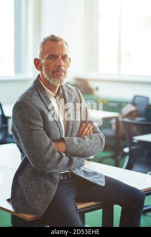 Uomo d'affari sicuro maturo in usura formale tenendo le braccia incrociate e. guardando la macchina fotografica mentre si appoggia sulla scrivania nel moderno ufficio o spazio di lavoro Foto Stock