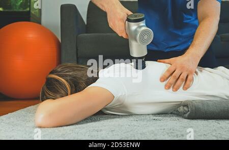 Terapista massaggiando la schiena della donna con un dispositivo di percussione massaggiante a casa sua. Foto Stock