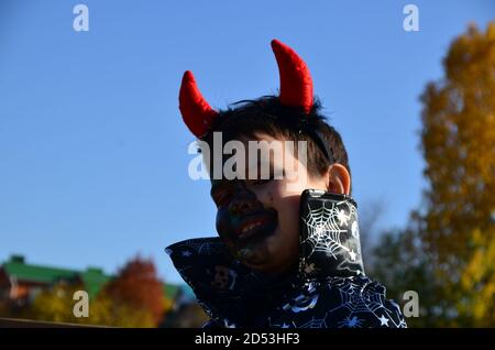 bambino divertente in costume halloween diavolo con corna e tridente su un ragazzo di sfondo di legno scuro con trucco nero per halloween, zombie Foto Stock