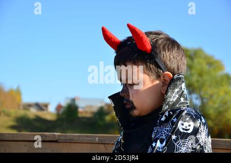 bambino divertente in costume halloween diavolo con corna e tridente su un ragazzo di sfondo di legno scuro con trucco nero per halloween, zombie Foto Stock