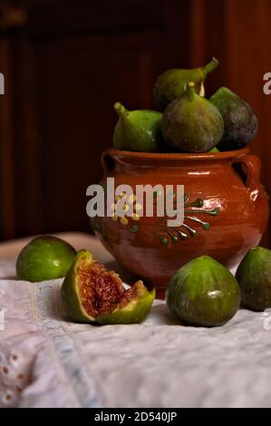 Un barattolo marrone pieno di fichi e qualche frutta intorno su un panno decorato e un fondo di legno duro di alimento scuro Foto Stock