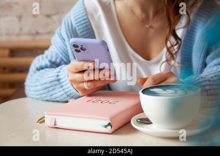 Giovane donna in pullover blu caldo sta leggendo un testo messaggio dal telefono cellulare mentre si beve caffè Foto Stock