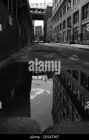 Riflessione gigante in puddle lungo Plymouth Street a DUMBO, Brooklyn, verso i ponti di Manhattan e Brooklyn. Strada deserta. Monocromatico, nero e wh Foto Stock