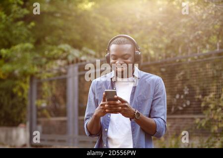 Passeggiate per la strada, relax, tempo libero. Giovane uomo afro-americano in cuffia che ascolta la musica sullo smartphone utilizzando l'app musicale. Ritratto di sorridere Foto Stock