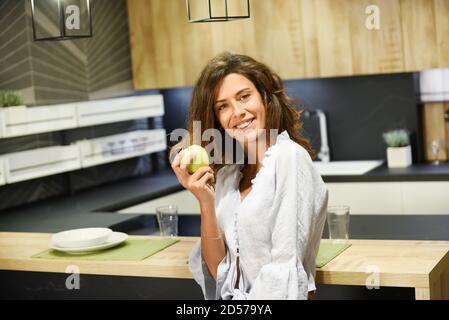 Donna vivace sorridente che tiene in mano una mela verde fresca cucina a pianta aperta in un concetto di dieta sana e nutrizione Foto Stock