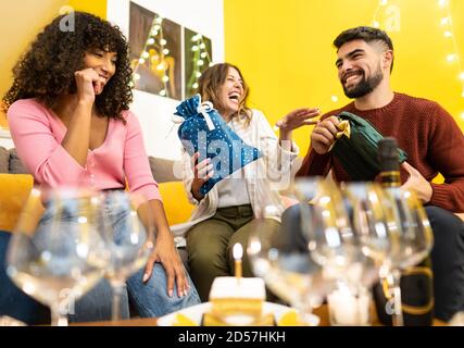 Due giovani e bei amici caucasici danno il loro dono amico nero che celebra il suo compleanno a casa ridendo e scherzando - gara mista a casa par Foto Stock