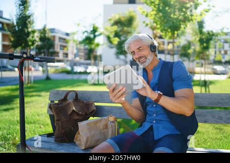 Ritratto di un uomo maturo con le cuffie che si trovano all'aperto in città, utilizzando un tablet. Foto Stock