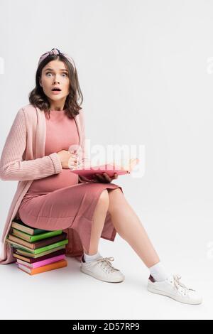 donna scioccata e incinta seduta, tenendo libro e toccando pancia su bianco Foto Stock