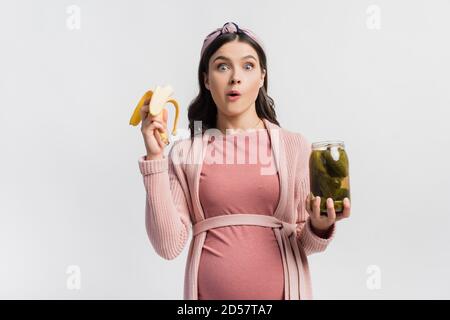 donna incinta che mangia banana mentre tiene il vaso con i cetrioli in scatola isolato su bianco Foto Stock