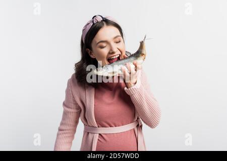 donna incinta con bocca aperta e occhi chiusi che tengono asciugato pesce isolato su bianco Foto Stock