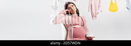 raccolto panoramico di donna pessiva e incinta che tiene minuscoli stivali mentre si siede vicino a vestiti del bambino isolati su bianco Foto Stock