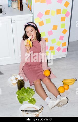 donna incinta che mangia banana vicino al vaso con cetrioli in scatola, cupcakes e arance mentre si siede sul pavimento Foto Stock