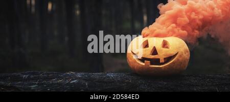 zucca di halloween con fumo arancione su tronco di albero in fondo foresta scuro. copia spazio Foto Stock