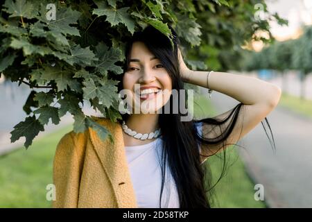 Giovane bella bruna asiatica allegra ragazza indossando elegante blazer giallo e tshirt bianca, in posa nella strada della città sotto l'albero d'acero, sorridente a. Foto Stock