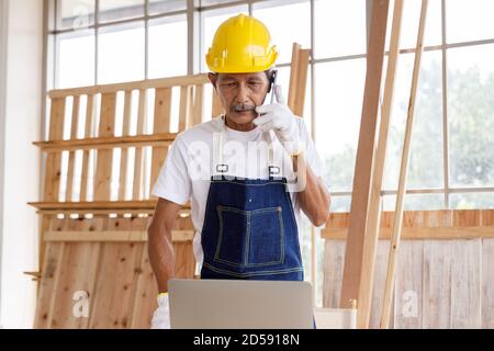 Ingegnere senior o falegname parlare al telefono e guardare il computer portatile in officina di Falegnameria. Foto Stock