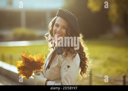 Ciao ottobre. Donna elegante sorridente in trench camice beige con foglie gialle autunnali fuori sulla strada della città in autunno. Foto Stock