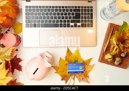 Ciao autunno. lay piatto con taccuino, foglie, castagne, banca piggy, carta di credito e laptop su tavolo bianco. Foto Stock