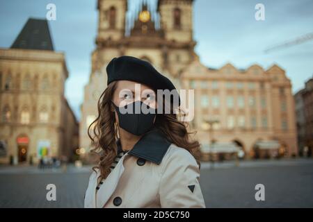 Ciao autunno. Moderna donna di mezza età in trench beige e beretto nero con maschera nera a Piazza della Città Vecchia di Praga. Foto Stock