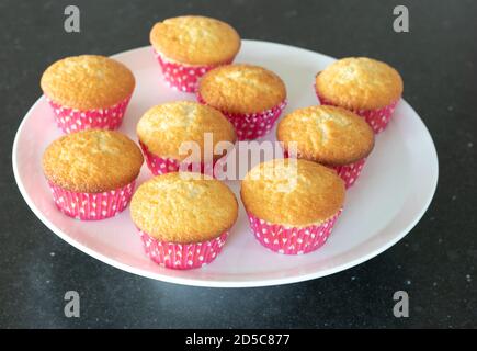 Plein cupcakes cotto in casa in un piatto bianco, vista dall'alto su uno sfondo grigio scuro, tazze rosa con puntini bianchi Foto Stock