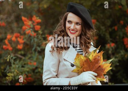 Ciao ottobre. Sorridente elegante donna di mezza età in trench beige con foglie gialle autunnali all'aperto in città in autunno. Foto Stock