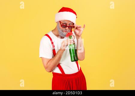 Divertente uomo con bearded grigio in costume di babbo natale bevendo bevande alcoliche da bottiglia con ombrello di carta, riposando nelle vacanze invernali. Monolocali interni Foto Stock