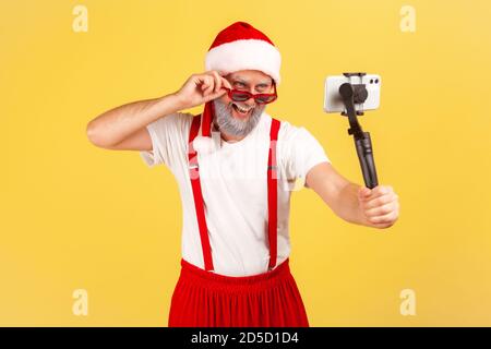 Bell'uomo grigio bearded in costume di babbo natale che tiene lo smartphone sul bastone in posa per fare foto o registrare video, blogging. Studio interno girato isolat Foto Stock