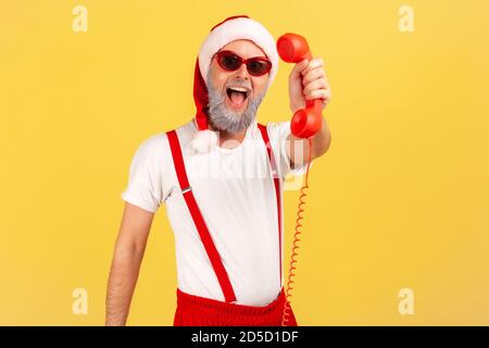 Eccitato felice uomo con bearded grigio in occhiali da sole e cappello di babbo natale che tiene e mostra telefono fisso, chiamando per congratularsi con le casse. IO Foto Stock