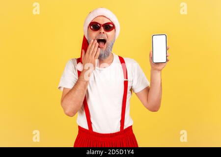 Uomo sorpreso scioccato in occhiali da sole eleganti e babbo natale rosso cappello che tiene smartphone con schermo vuoto, incredibili proposte online. Stu. Interno Foto Stock