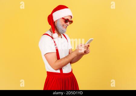 Profilo ritratto uomo felice in occhiali da sole e costume di babbo natale che chattano su smartphone, nessun traffico internet in messaggeri. Isolamento delle riprese in studio al coperto Foto Stock