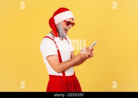 Felice uomo divertente in costume di babbo natale e occhiali da sole alla moda digitando sullo smartphone e sorridendo, leggendo piacevoli notizie. Studio al coperto isolato Foto Stock