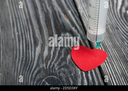 Un cuore di carta forato con una siringa medica. Le tavole di pino sono nere. Foto Stock